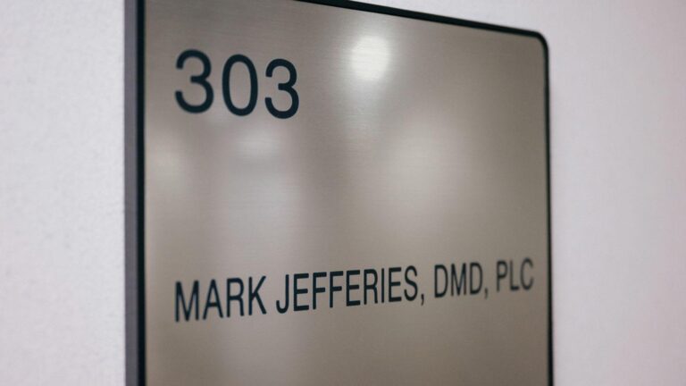 The image shows a metallic office sign mounted on a wall. The sign has the number "303" at the top and the name "MARK JEFFERIES, DMD, PLC" below it. This image is relevant as it indicates the office location and the name of the professional, likely a dentist, working in room 303.