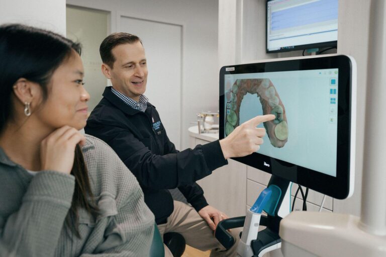 The image shows a dental professional pointing at a computer screen displaying a 3D model of a dental arch. The screen is part of a dental imaging system, likely used for planning orthodontic treatments or other dental procedures.