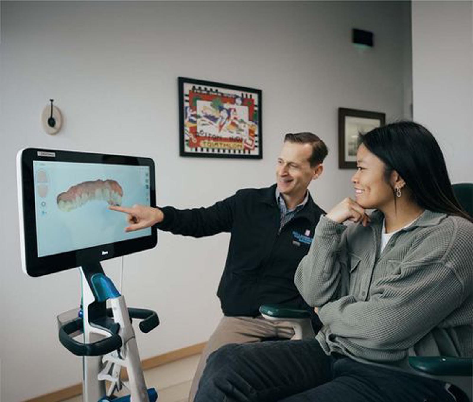 The image shows two individuals in what appears to be a dental office. One person is pointing at a computer screen displaying a 3D model of teeth, while the other individual is seated, attentively observing. The setting includes framed artwork on the walls and dental equipment, reinforcing the professional environment.