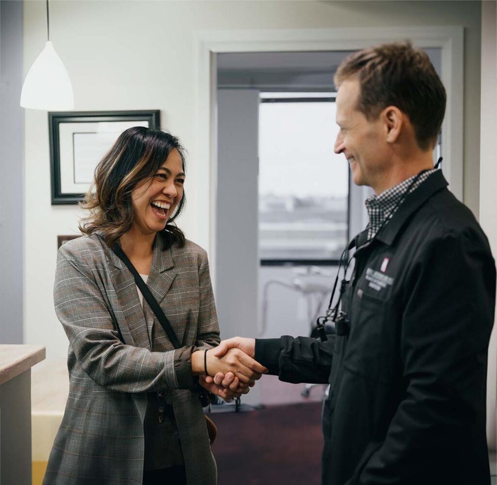 The image captures a handshake between two individuals in what appears to be a professional or medical setting. One person is wearing a plaid blazer and carrying a shoulder bag, while the other is dressed in a dark jacket with a name tag and a checkered shirt underneath.