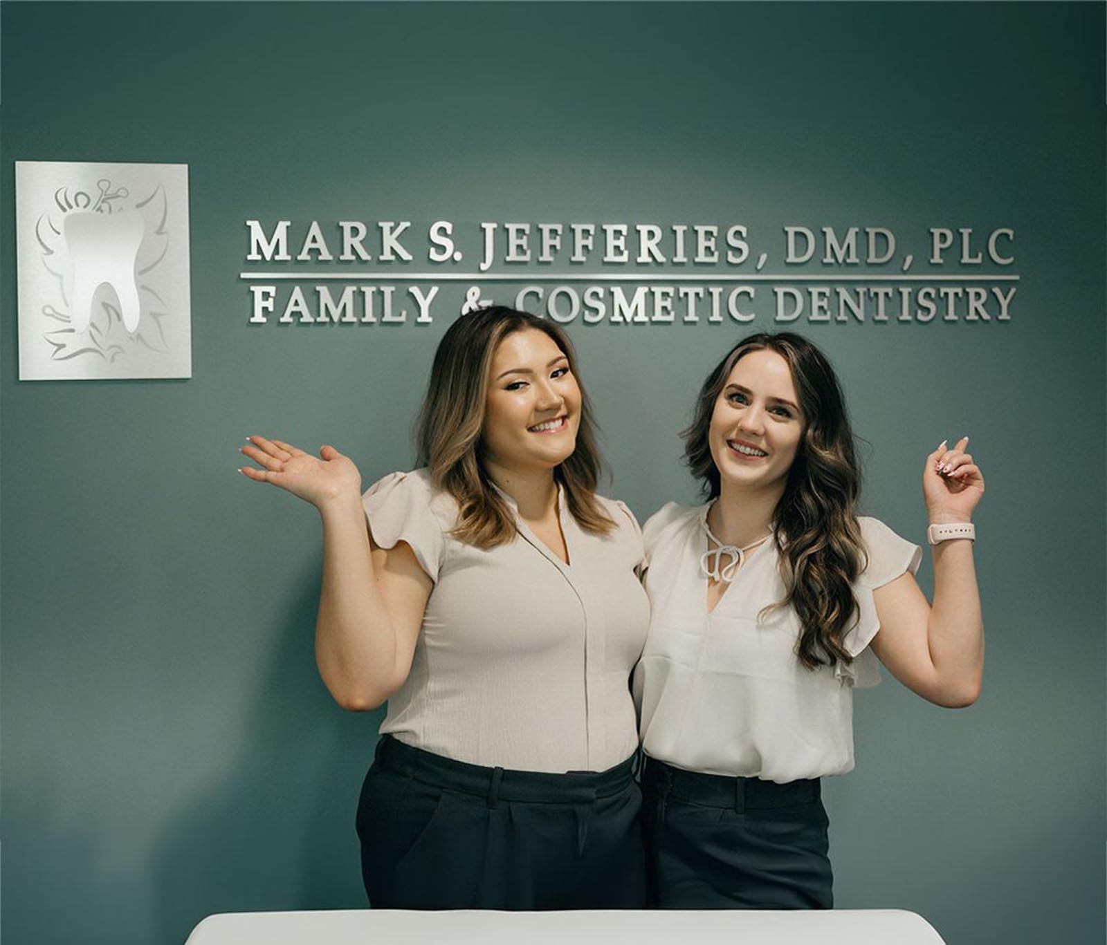 The image displays two individuals standing in front of a wall sign that reads "MARK S. JEFFERIES, DMD, PLC FAMILY & COSMETIC DENTISTRY." A stylized tooth logo with decorative elements accompanies the text. Both individuals are dressed in professional attire, with light-colored tops and dark pants, reinforcing the formal setting.
